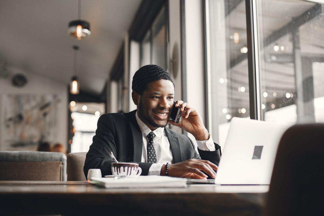 Smiling businessman in suit multitasking on laptop and phone at café.